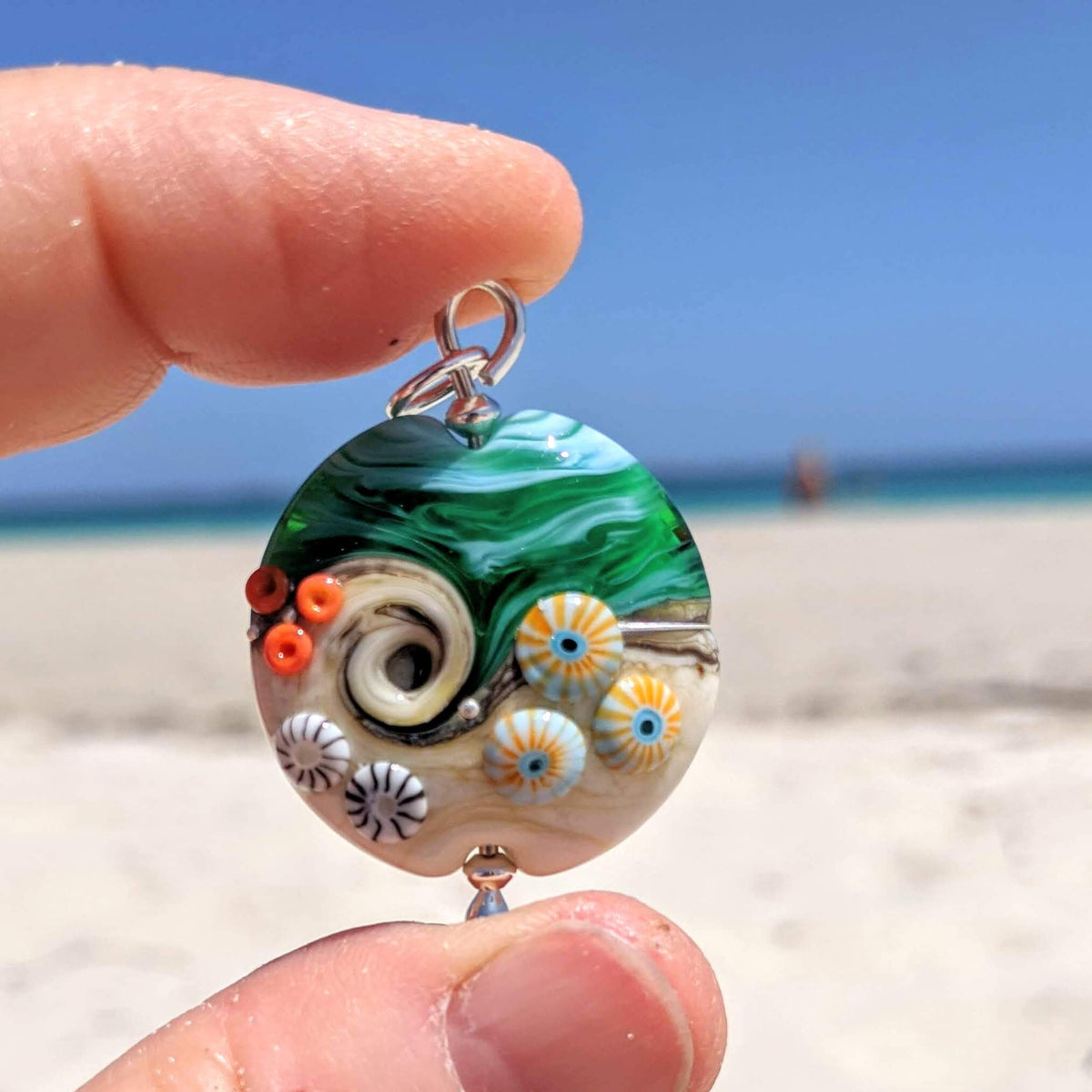 Pendant held up against a blue sky and white sand beach