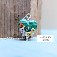Heart-shaped pendant with beachy designs on a wooden surface, branded 'Sand & Sea Classic'.